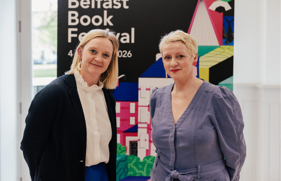 Two women standing in front of a banner and smiling at the camera. The banner reads 'Belfast Book Festival'.