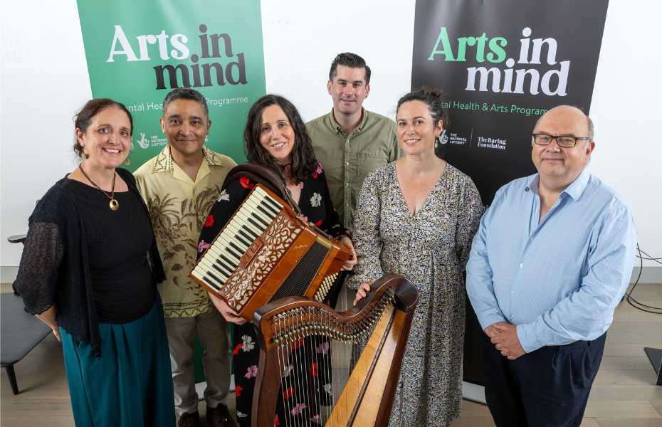 A group of three women and three men standing in front of two bannerstands which say Art in Mind. They are looking at the camera and smiling and the three women are holding instruments.