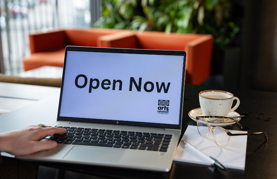 An open laptop with the words Open Now on the screen. A pair of glasses and a coffee cup are sitting beside it.