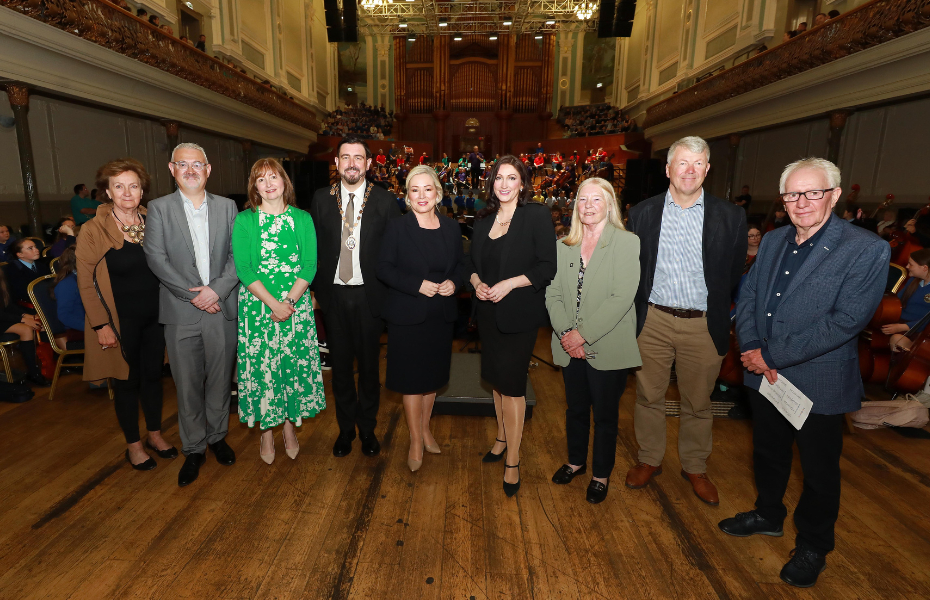 The group standing inside the Ulster Hall smiling at the camera with the Ulster Orchestra and young people on the stage in the background.
