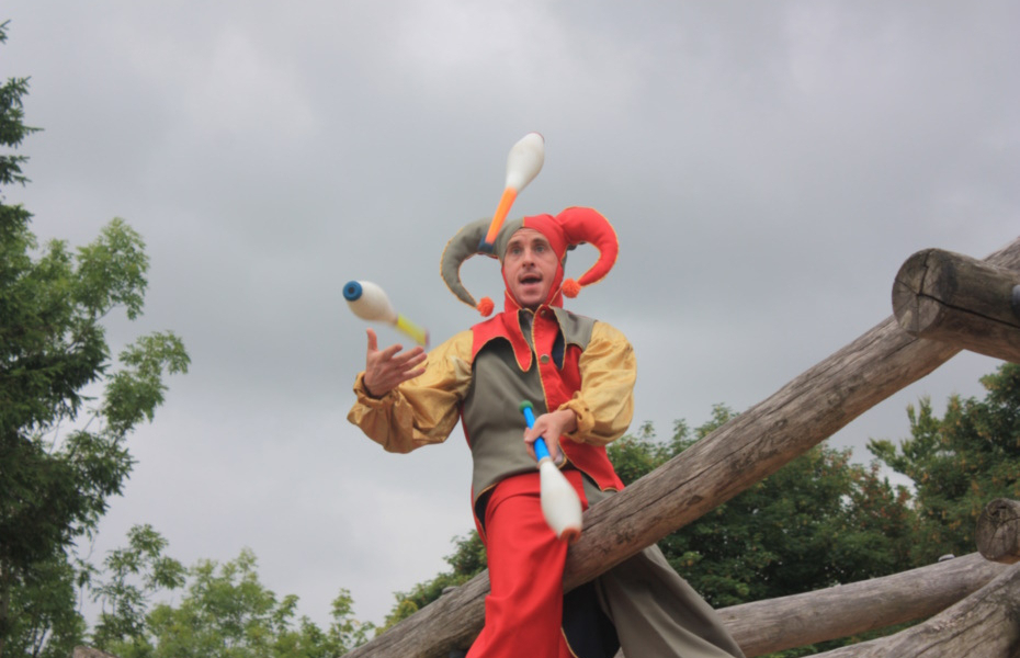 An In Your Space Circus artist juggles bowling pins. There are trees and grey skies in the background, making their colourful outfit stand out.