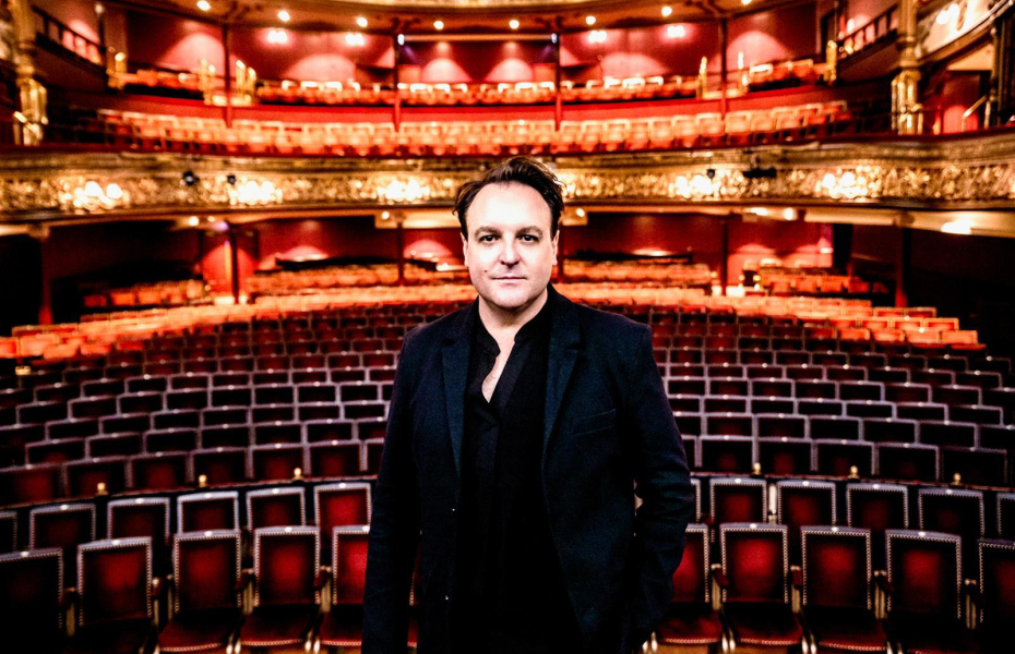 A man in a black shirt and suit on stage at the Grand Opera House, with lights on and theatre seating behind him.