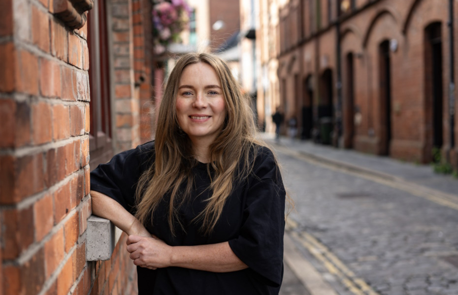Claire Kieran standing outdoors against a red brick wall wearing a black t-shirt and smiling towards the camera.