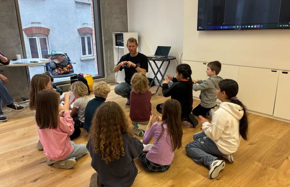 A group of young children sat on the floor in a room using sign-language looking at a trainer who is also using sign-language.