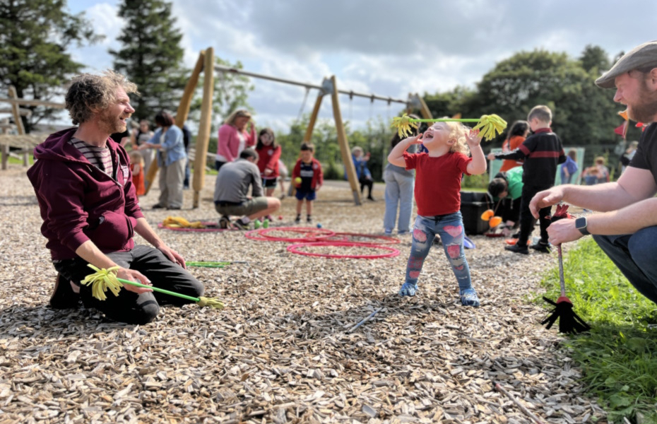 A young child throwing a stick in the air with a huge smile on their face.  The child is outside in a park area with swings in the background and people playing.