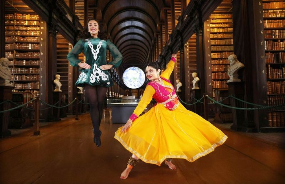 An Indian lady dancer in a bright yellow Indian dress, and an lady Irish dancer, with Trinity College Library in the background.