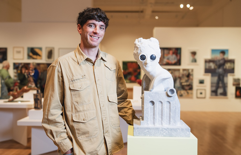 John is pictured in a gallery, artworks can be seen behind him. The white bust of a head has black goggles on its eyes