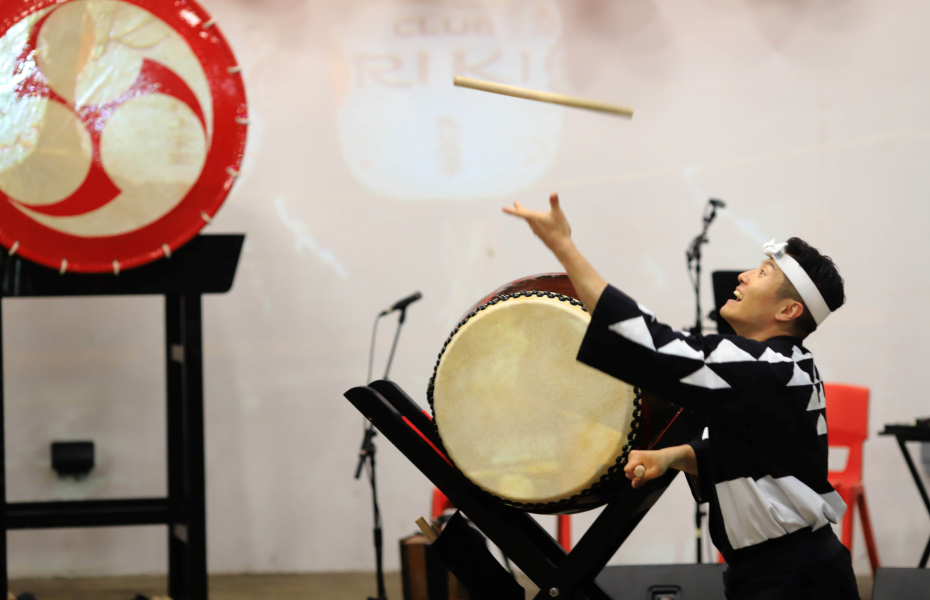 An image of a drummer, he throws a drum stick in the air, behind him are large Japanese drums