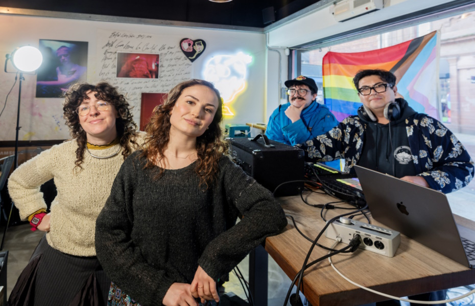 Four people standing beside musical equipment with an LGBTQI+ flag in the background.