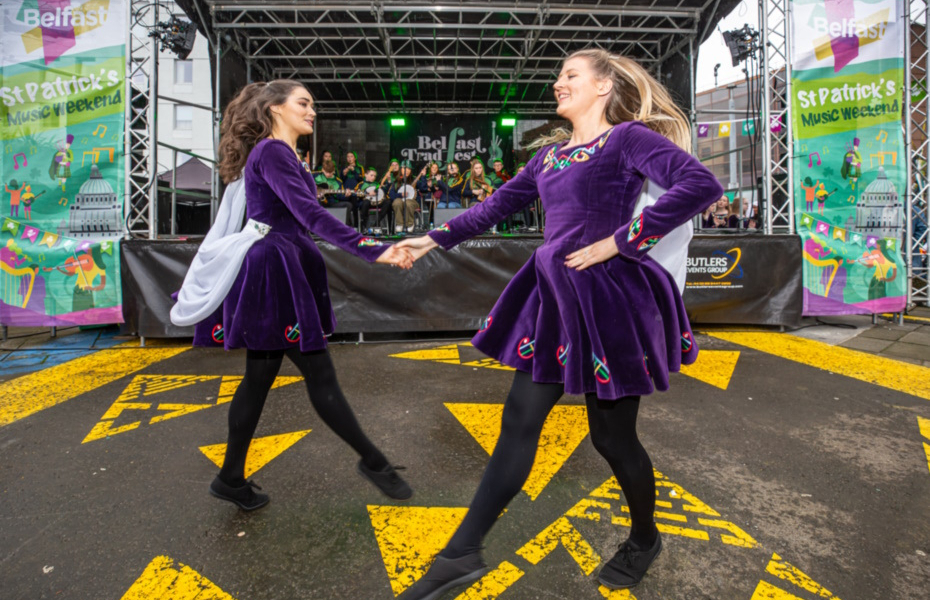 Two Irish Dancers in purple velvet costume dancing beside a stage.