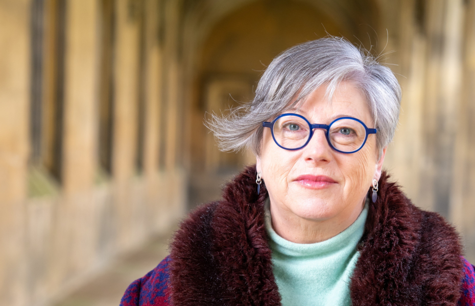 A woman with short grey hair, wearing glasses and a purple coat, facing the camera, with pillars of a university in the background.