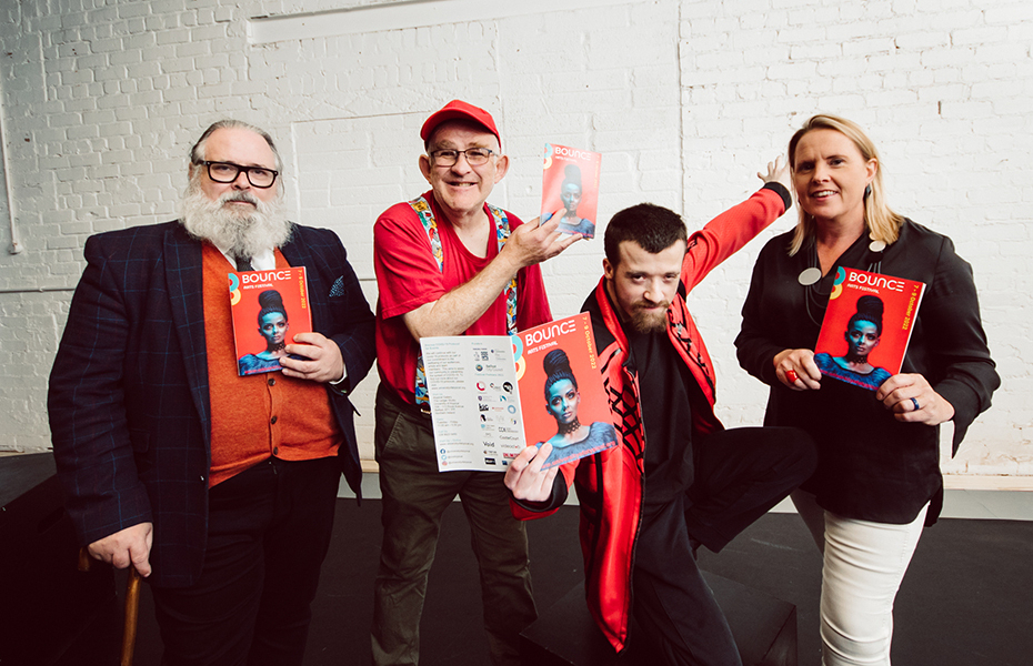 Three males and one female standing against a white stone background holding the printed programme for the 2022 Bounce! Festival.