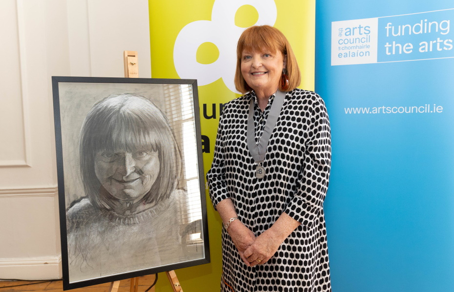Image of a woman smiling, with silver Laureate na nÓg medal around her neck, pictured in front of a black and white portrait of her on a large easel.
