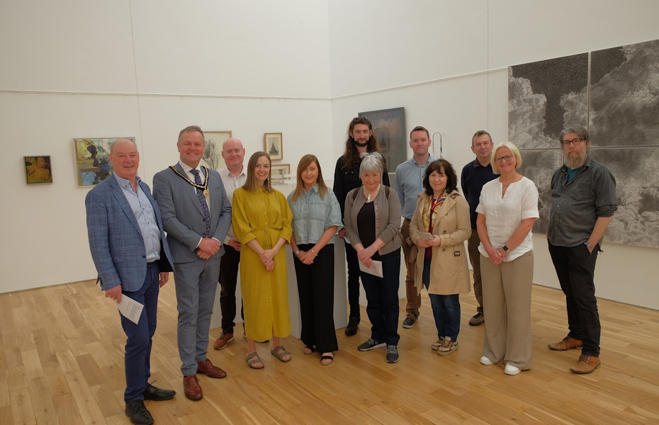 Image of a large group in a white gallery. Behind them you can see artworks displayed on the walls. They are all smiling.