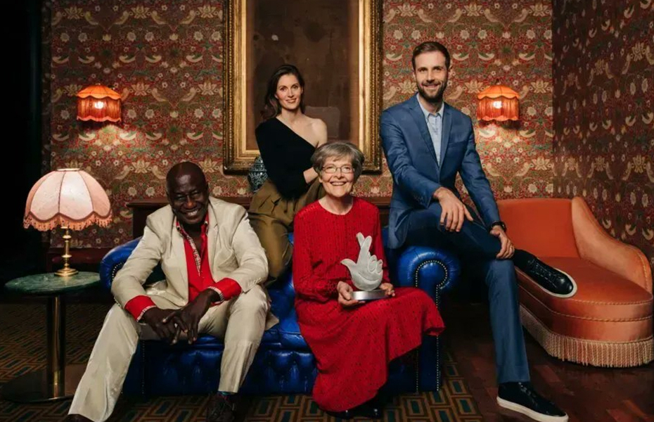 An image of four people sitting on a sofa in a room with colourful wallpaper. One person is holding a national lottery trophy.
