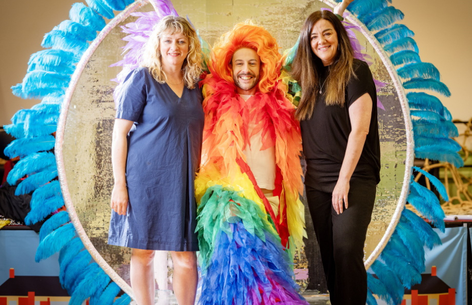 Three people stand, smiling, against a sparkling silver backdrop, encircled with blue feathers.