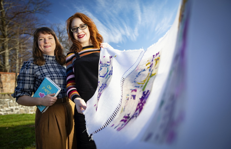 Two artists are pictured, one holds a book under their arm, the other a long piece of embroidered fabric which stretches towards the camera