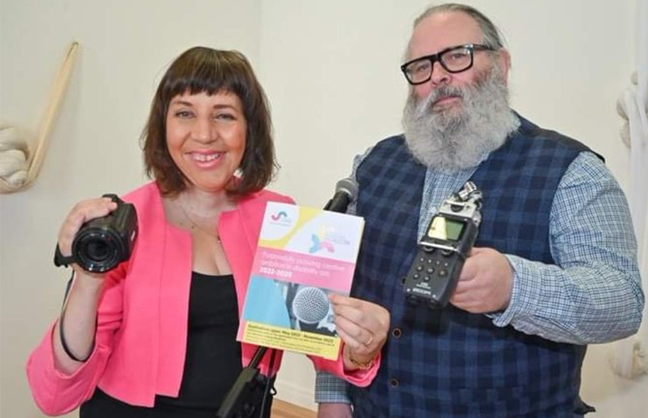 A female in a pink blazer and a male in a blue checked waistcoat holding a selection of digital equipment including a camcorder and a dictaphone.