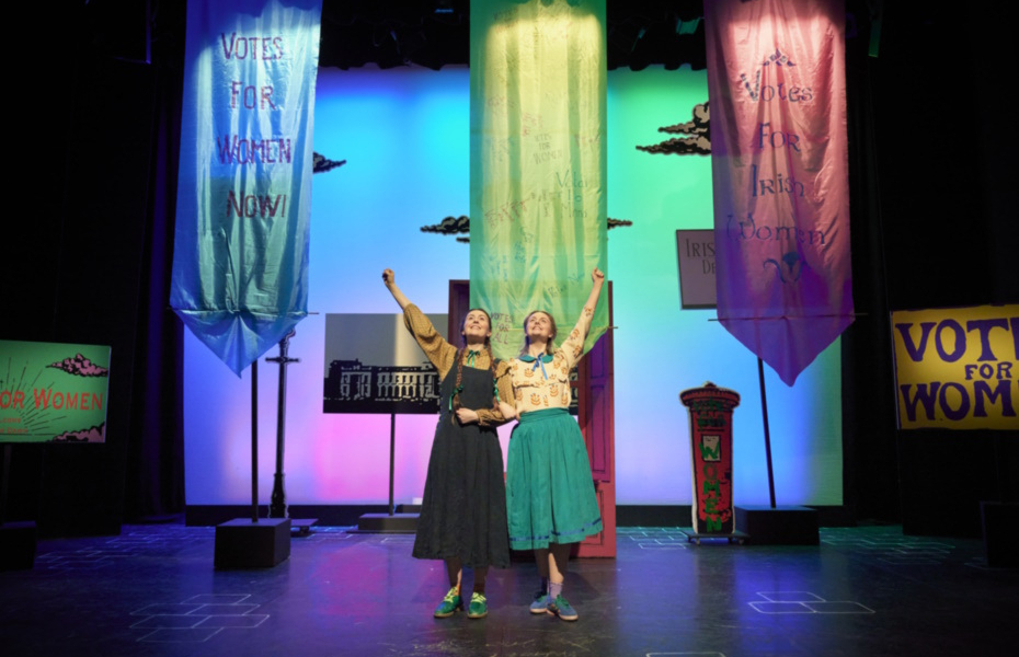 Two actors on stage with Votes for Women signage in the background.